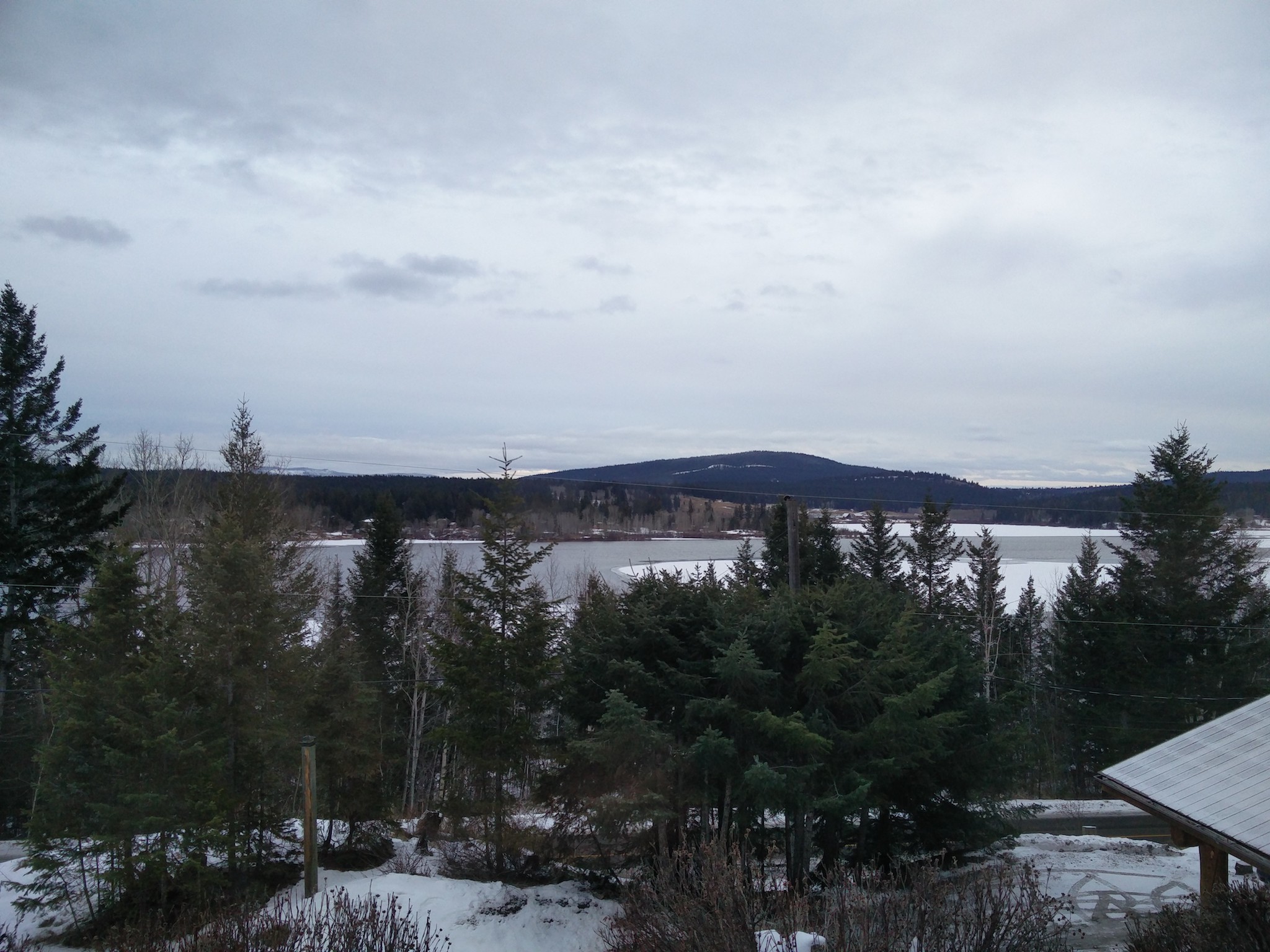 Par une fenêtre, on voit un lac glacé entouré de forêt et de montagnes. Au premier plan, vue plongeante vers la route blanche depuis la maison en hauteur.